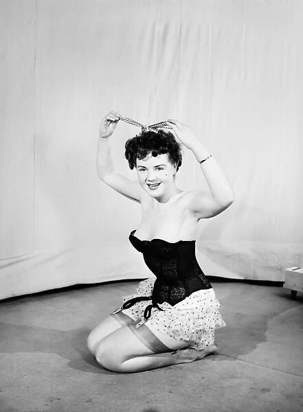Clothing: Fashion: Underwear: Woman wearing corsett placing bow in hair. 1959