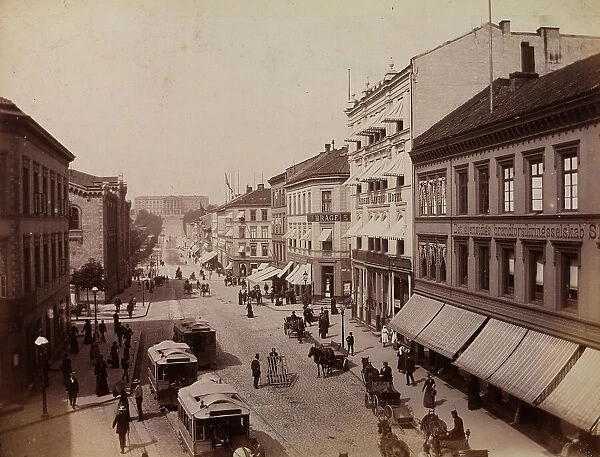Carl Johan Street in Oslo. A few wagons, horse drawn trams and people are passing. At the back of the street the Royal Palace can be glimpsed